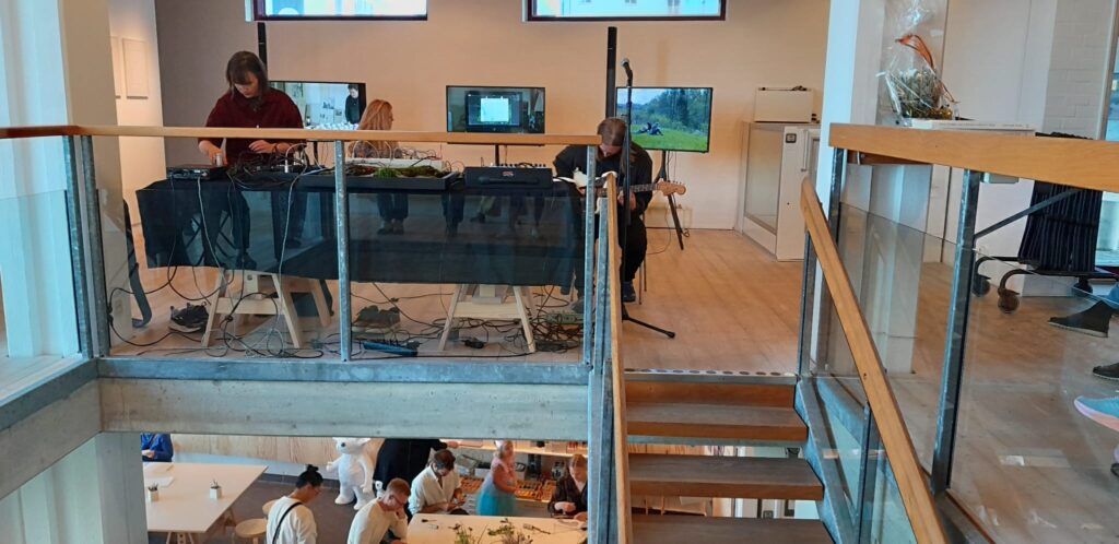 Två personer vid bord med ljudutrustning; bakom dem tre skärmar med filmklipp från natur och vatten.