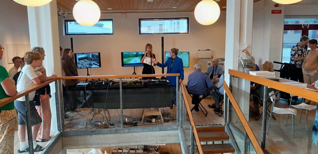 Talare presenterar utställningen framför publik; tre skärmar visar filmer från restaureringsarbete vid vattendrag.